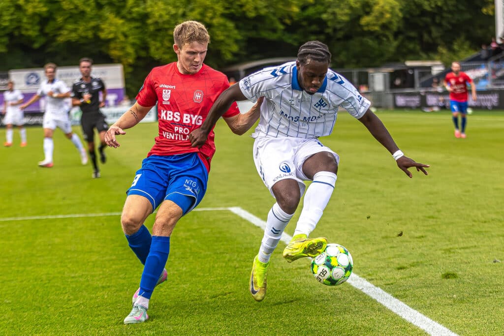 Intens duel om fodbolden i en kamp mellem Kolding IF i hvide og blå spilledragter og Hvidovre i røde trøjer og blå shorts på grønsværen