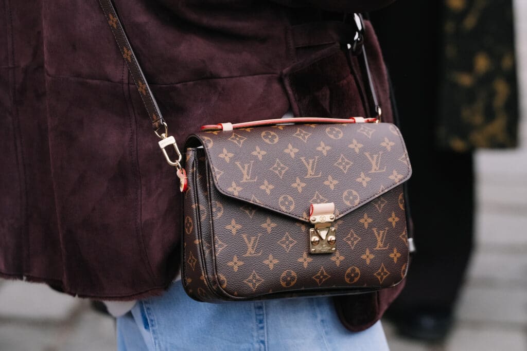 En klassisk Louis Vuitton Metis skuldertaske i brunt monogram-mønster med guldspænde båret cross-body over en mørkerød ruskindjakke